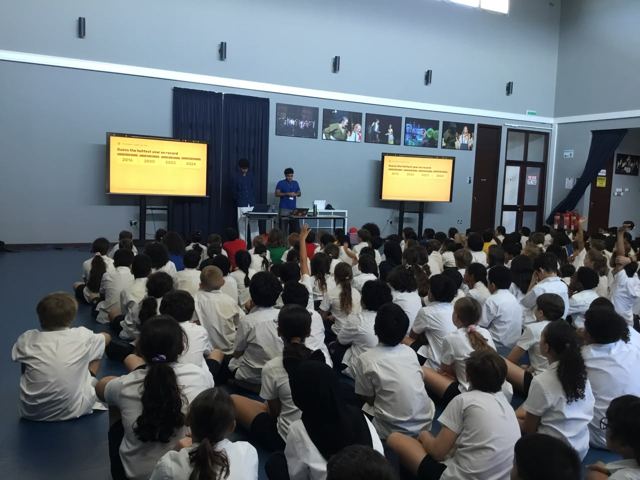 Students sit on the floor of a school hall facing two screens that ask them to guess the hottest year on record, with some hands raised.