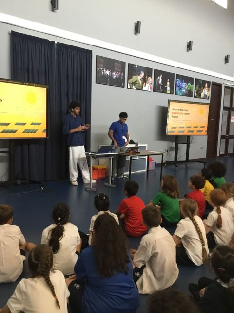Two presenters stand beside a table of laptops in front of seated students, with a screen showing the sun above solar panels.