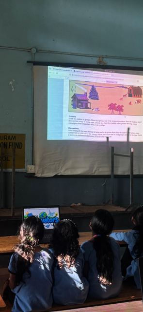 Students sit at wooden benches watching a projected lesson on energy, with a laptop showing the Gridovators logo.